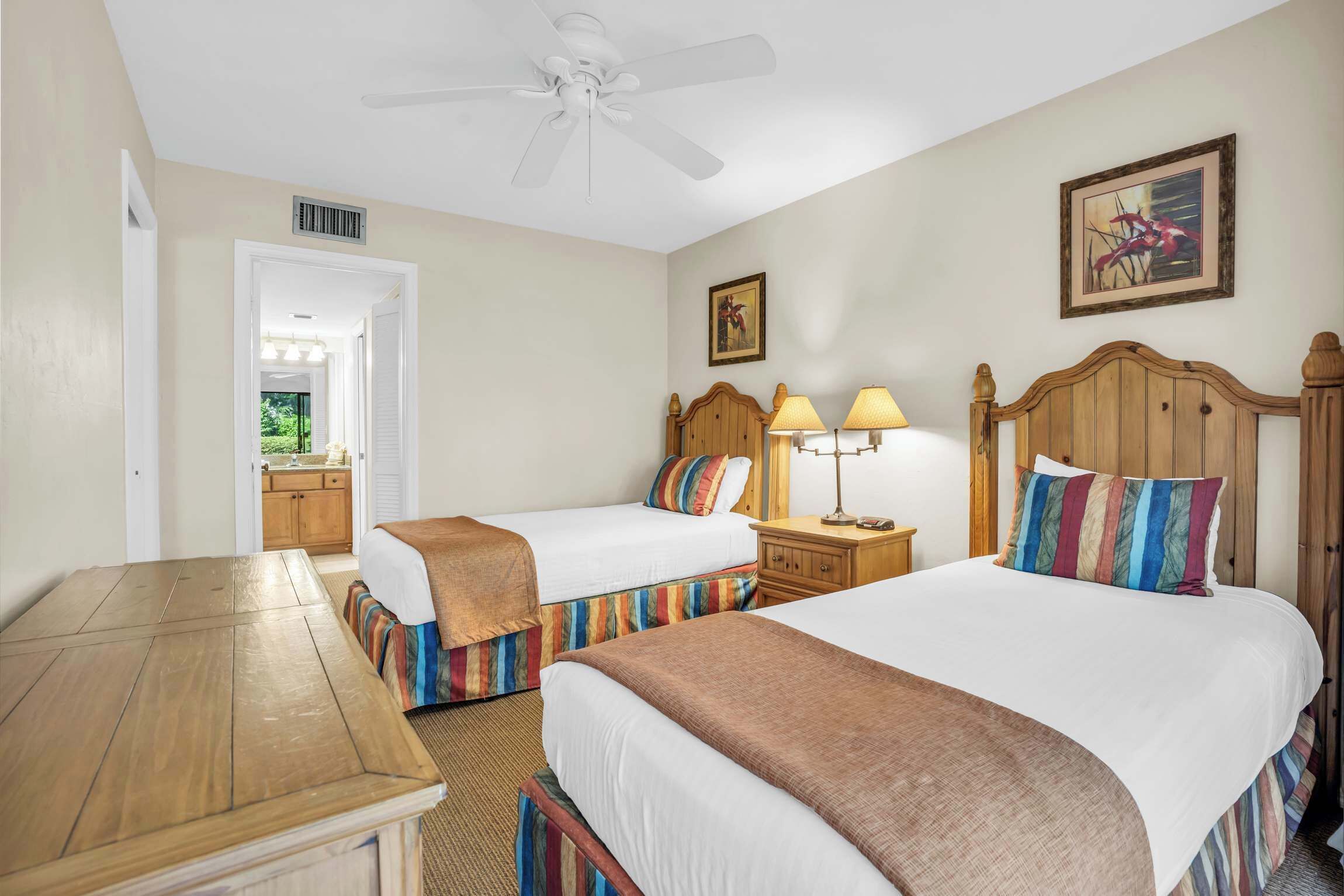 A hotel room with two beds, a wooden nightstand, a ceiling fan, and a painting on the wall.