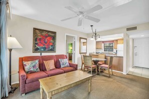 Un salon avec un canapé, une table basse et un tableau. On y trouve également un coin repas avec des chaises et une table. Une cuisine est visible au fond.