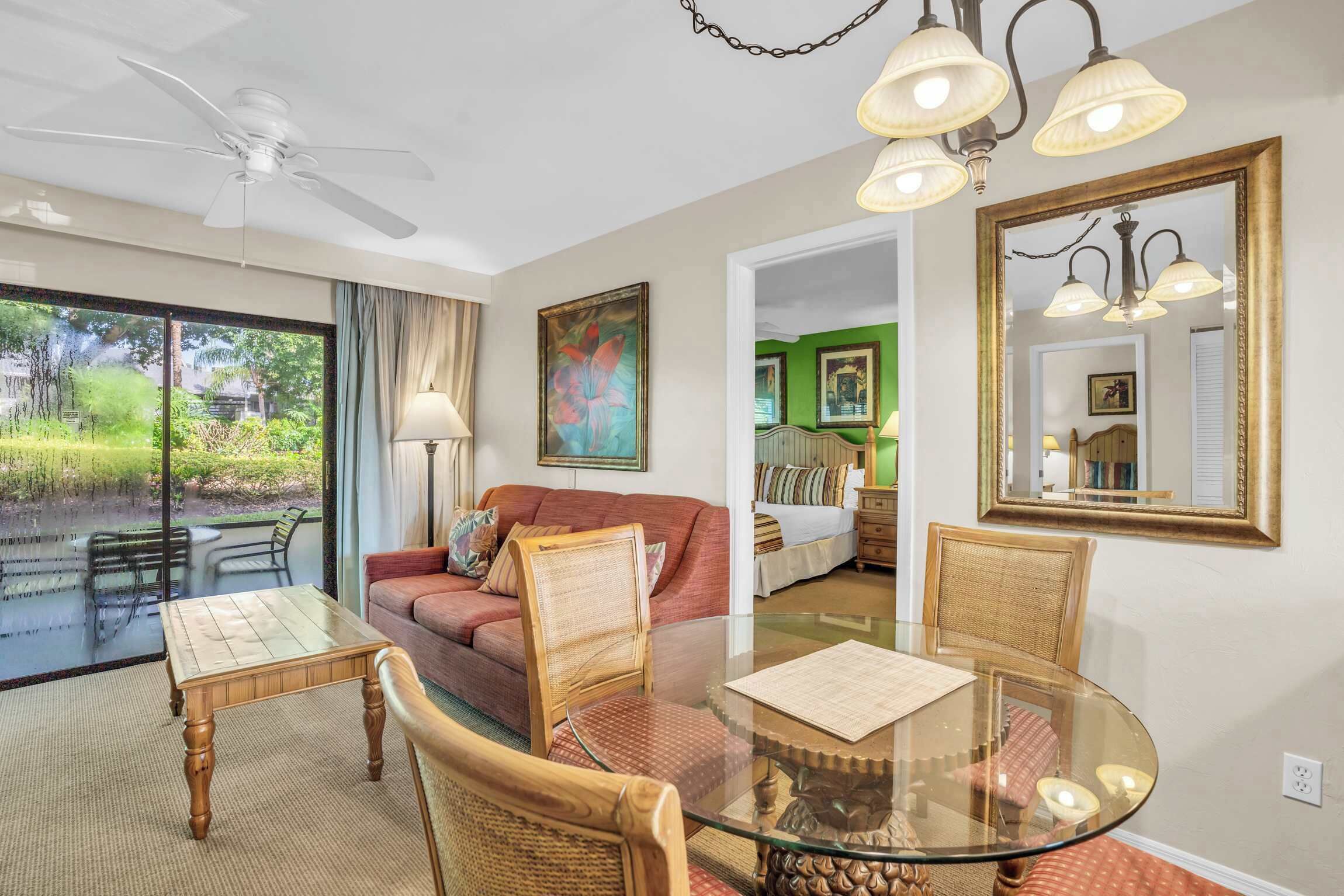 A room with a glass dining table, wicker chairs, a red sofa, a ceiling fan, and a large mirror.