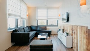 A modern living room with a sectional sofa, a small table, and a cabinet. There are large windows with blinds, a flat-screen TV mounted on the wall, and a wooden panel on one side.