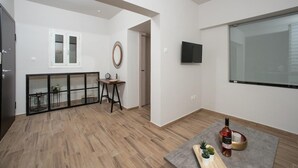 A modern living room with a wooden floor, a flat-screen TV mounted on the wall, a small table with a bottle and decorative items, and a window with a view.