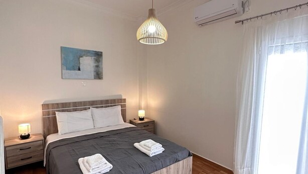 A modern bedroom with a wooden floor, a bed with grey bedding, a hanging light fixture, and a painting on the wall. - Nature House - At093 (Athens)
