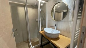 A bathroom with a glass-enclosed shower, a round mirror, and a wooden vanity with a white sink. - Eunoia Apartment - At062 (Athens)