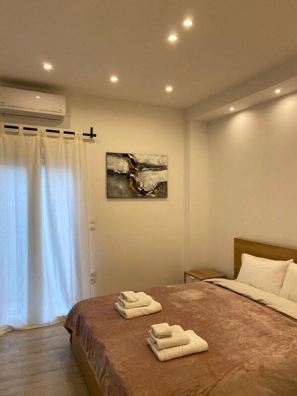 Een hotelkamer met een bed, nachtkastje, airconditioningapparaat, raam met gordijnen en een abstract schilderij aan de muur.