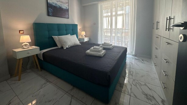 Un dormitorio con una cama grande, dos mesitas de noche con lámparas, una ventana con cortinas translúcidas y un armario con varios cajones.