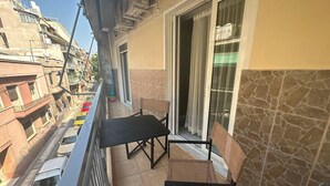 Un balcón con dos sillas negras y una mesa, una puerta corrediza de vidrio y vista a una calle concurrida con coches aparcados.