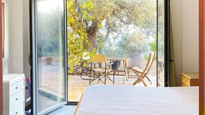 Une chambre avec un lit, un ventilateur de plafond et une porte-fenêtre coulissante donnant sur une terrasse extérieure équipée d’une table, de chaises et d’un arbre.