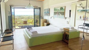 Une chambre avec un grand lit, un plancher en bois, un ventilateur de plafond et une vue sur l’extérieur, visible à travers une porte vitrée.