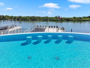 A clear blue pool with six cylindrical objects in the center, surrounded by lounge chairs and a deck with a railing.