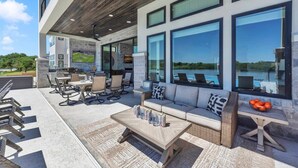 A modern outdoor patio with a stone fireplace, seating area, and large windows overlooking a body of water. - Lone Star Landing II | Concierge Services (Kingsland)