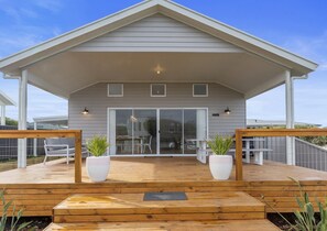 Eine überdachte Terrasse mit Holzboden, zwei Pflanzkübeln und einer Glastür, die zu einem Innenbereich mit Tisch und Stühlen führt.
