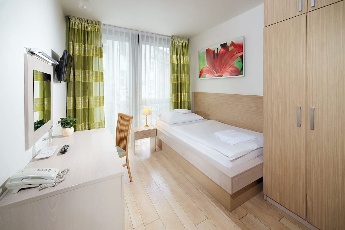 A hotel room with a bed, a desk with a telephone, a chair, a bedside table with a lamp, and a wardrobe.