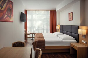 Quarto de hotel com cama, mesa de cabeceira, luminária, cadeira e televisão na parede.