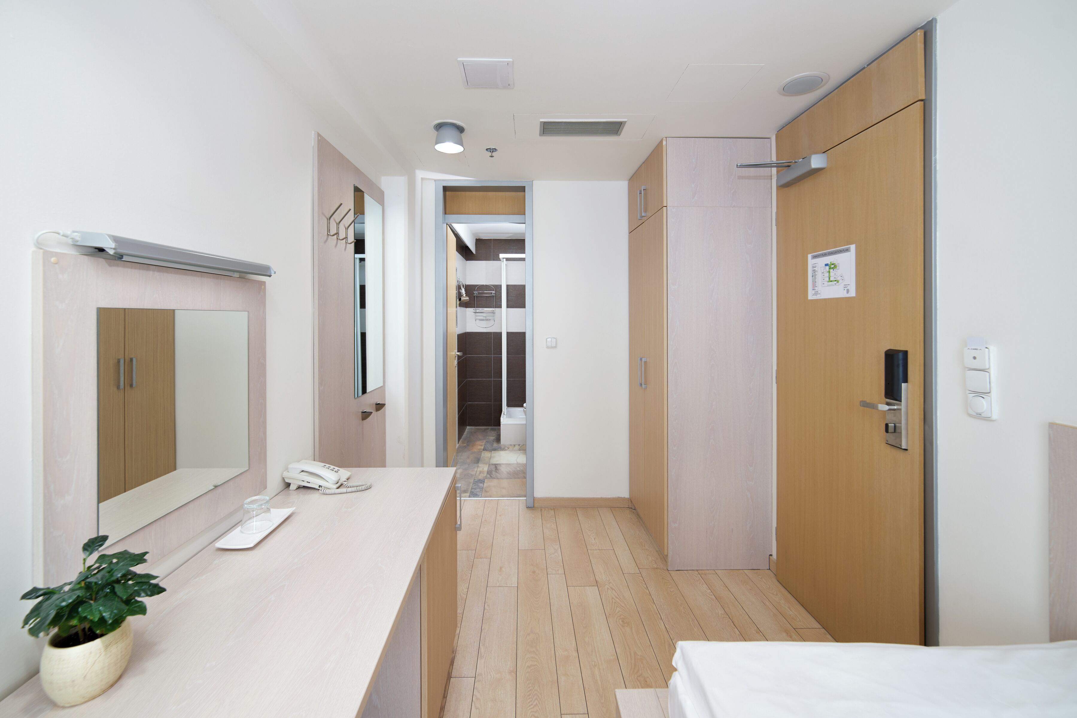 Habitación de hotel con piso de madera, mesita de noche con teléfono y vaso, espejo y puerta con un letrero.