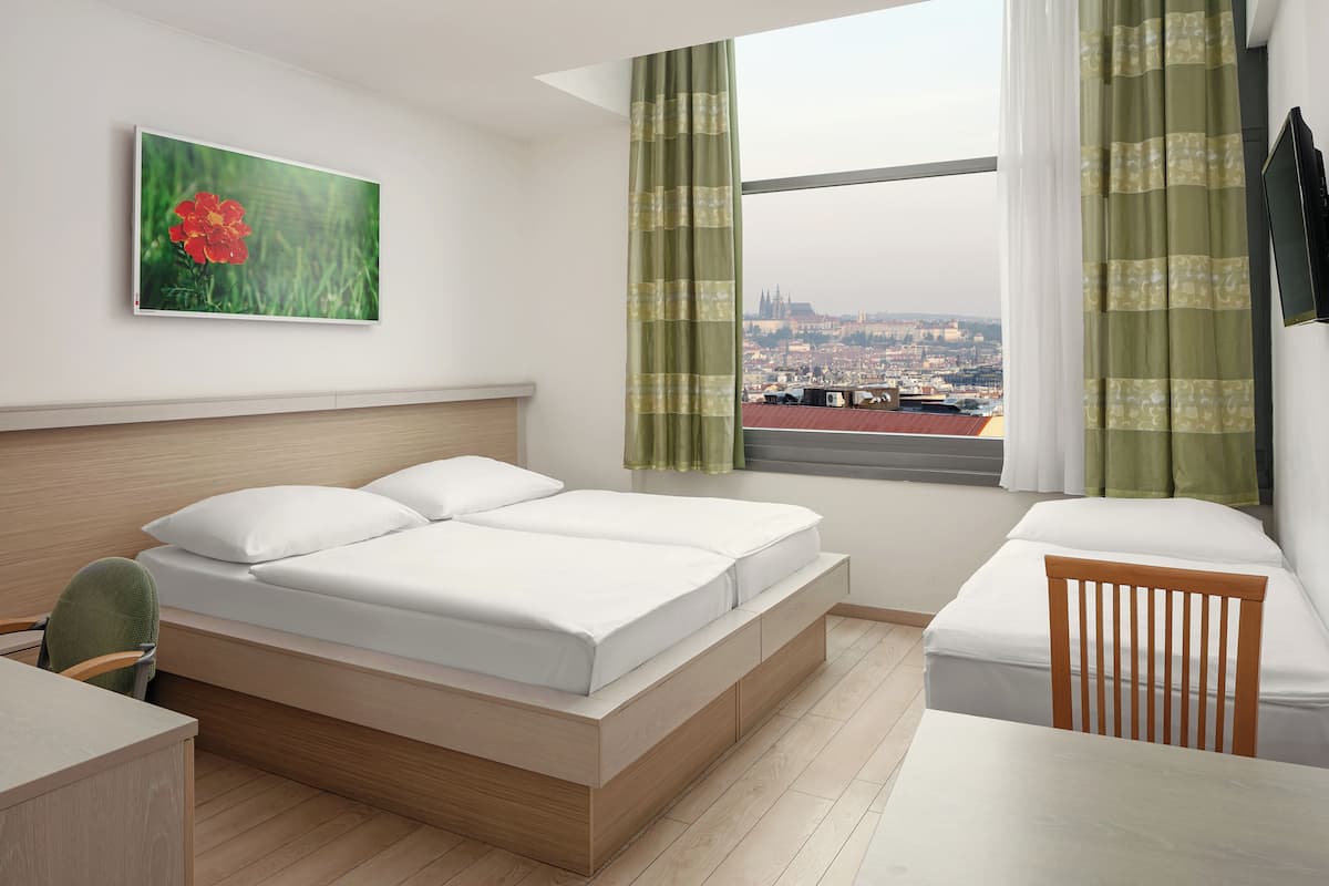 A hotel room with a bed, a desk, a chair, a window with a view of a cityscape, and a painting on the wall.