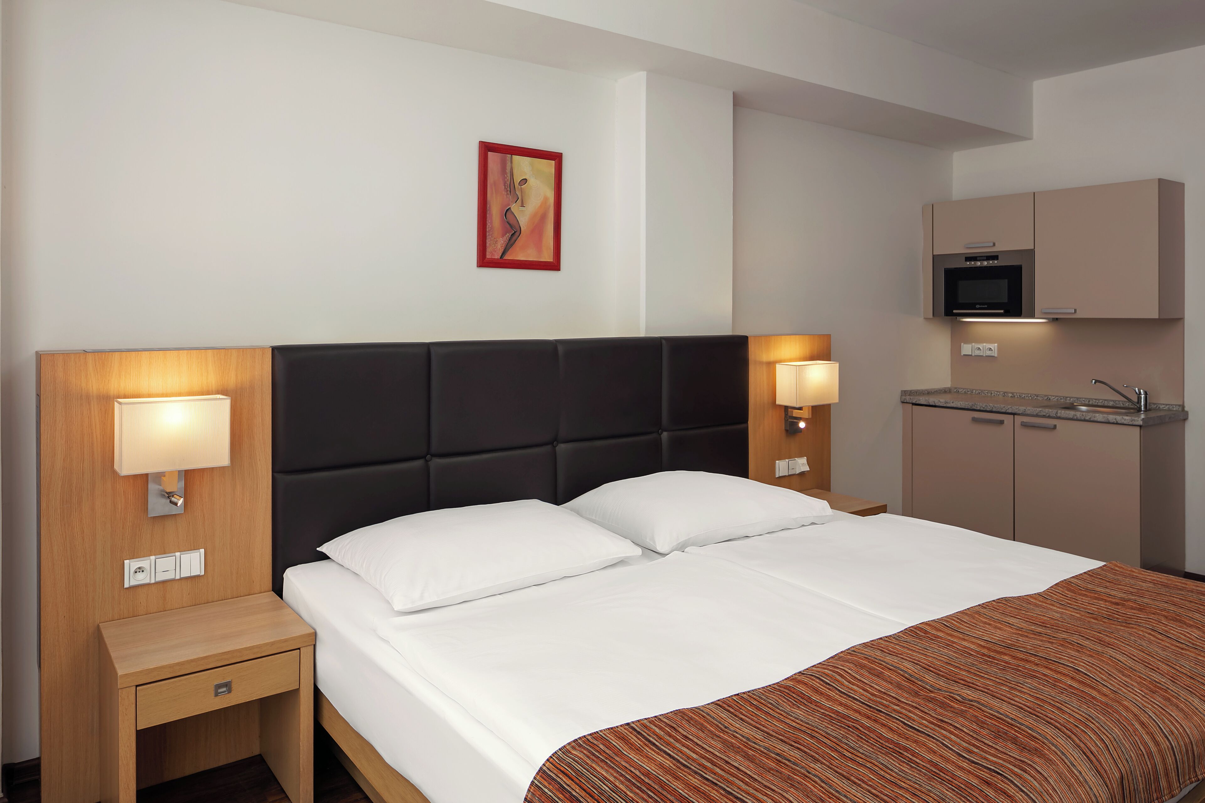 A hotel room with a bed, bedside lamps, a small table, a nightstand, a wall-mounted artwork, and a kitchenette.