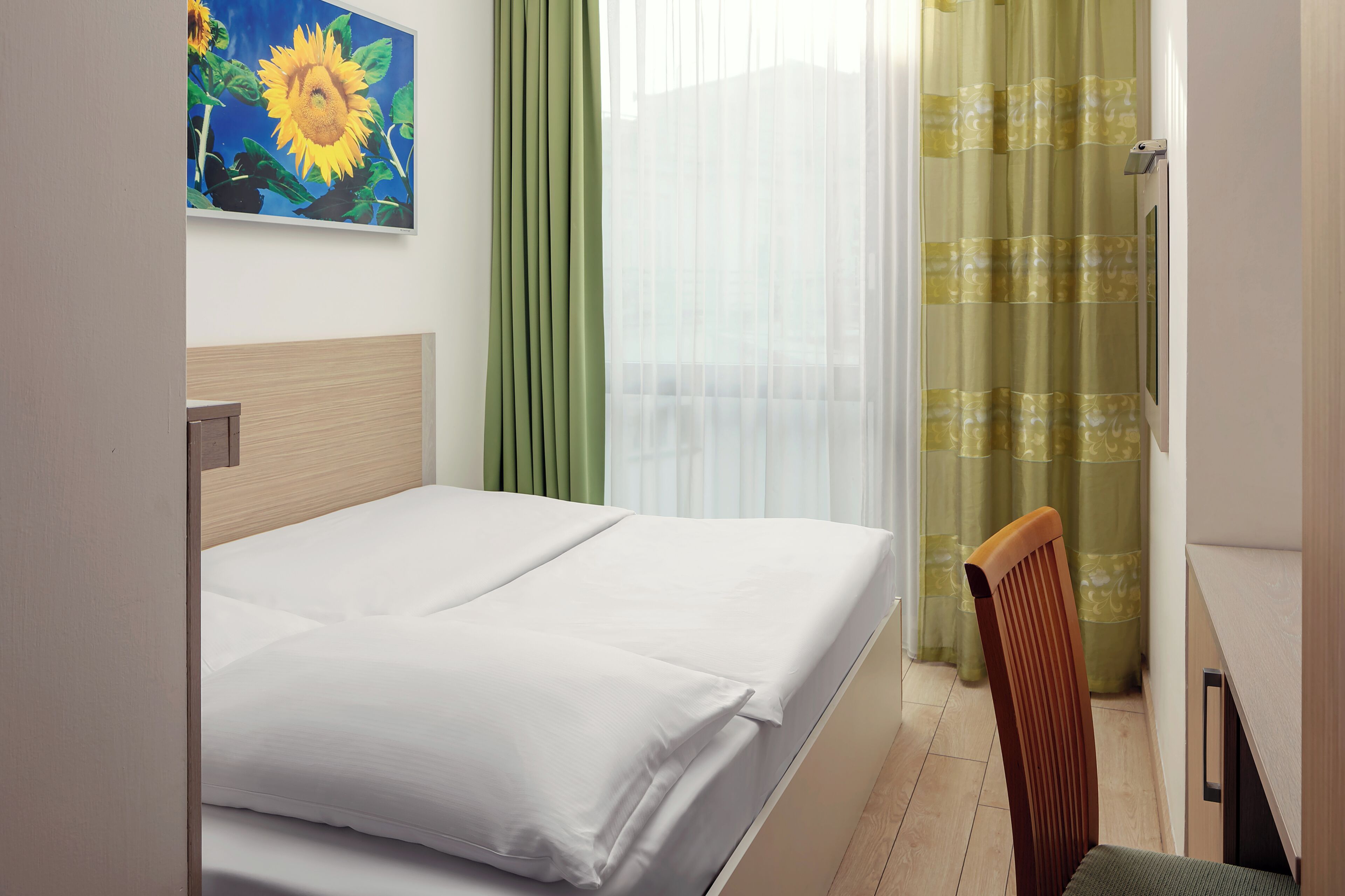 Habitación de hotel con cama, una silla, un cuadro de un girasol y vista al exterior a través de cortinas traslúcidas.