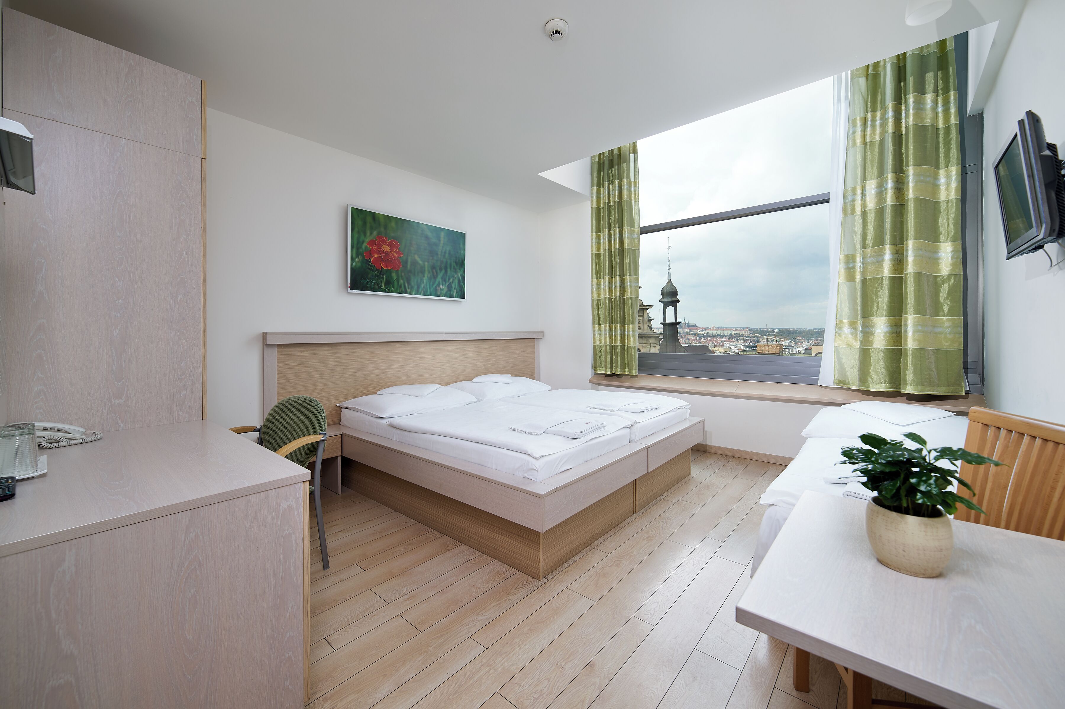 A modern hotel room with a bed, desk, chair, and a view of a cityscape through the window.