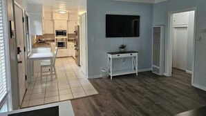 Uma sala de estar moderna com uma televisão de tela plana, um aparador branco e uma cozinha ao fundo.