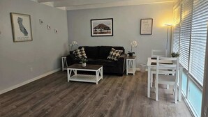 Sala de estar com sofá preto, mesa de centro branca e área de jantar com cadeiras brancas. Há quadros nas paredes e uma janela com persianas.