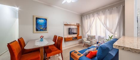 A modern living room with a dining area, a ceiling fan, a TV mounted on a wooden panel, and a large window with sheer curtains.