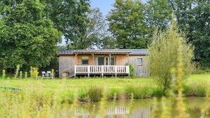 Une cabane en bois dotée d’un porche, entourée d’un étang et d’arbres.