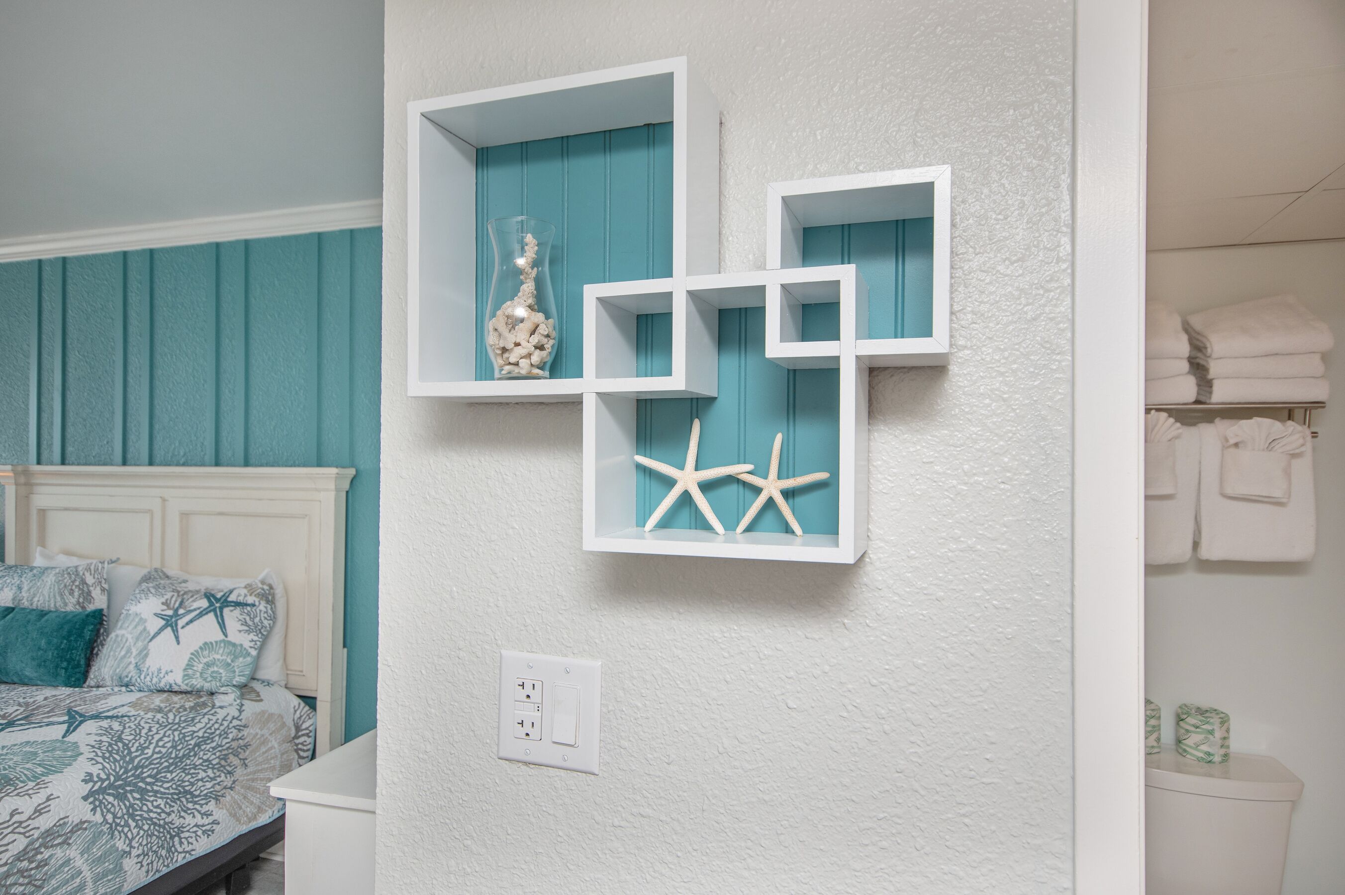 A bedroom with a bed, a wall-mounted shelf with decorative items, and a white wall switch.