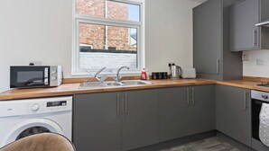 Une cuisine compacte avec des meubles gris, un lave-linge, un four à micro-ondes et une fenêtre donnant sur un immeuble en briques.