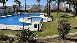 En swimmingpool omgiven av ett staket, med palmer och en byggnadskomplex i bakgrunden.