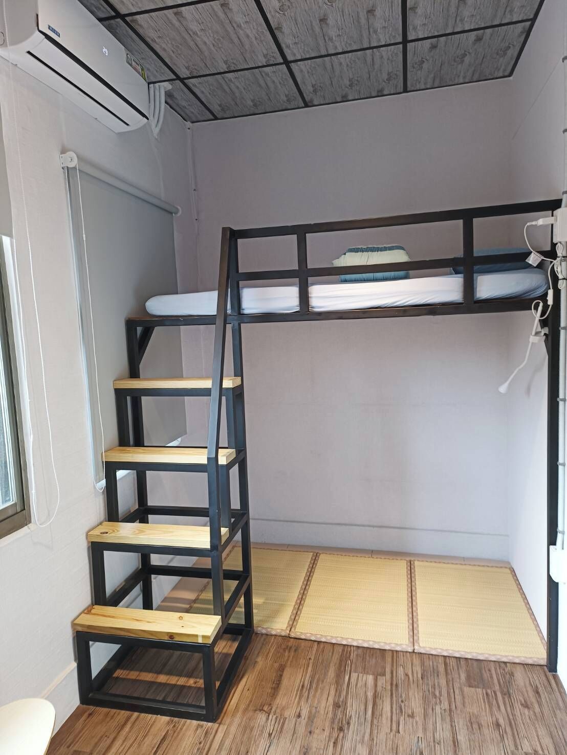 A bunk bed with a wooden ladder, a window, and a mat on the floor.