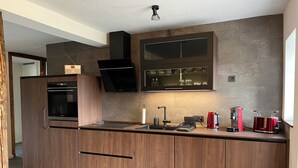 Una cocina moderna con armarios de madera, horno empotrado, pileta de acero inoxidable y tetera roja.
