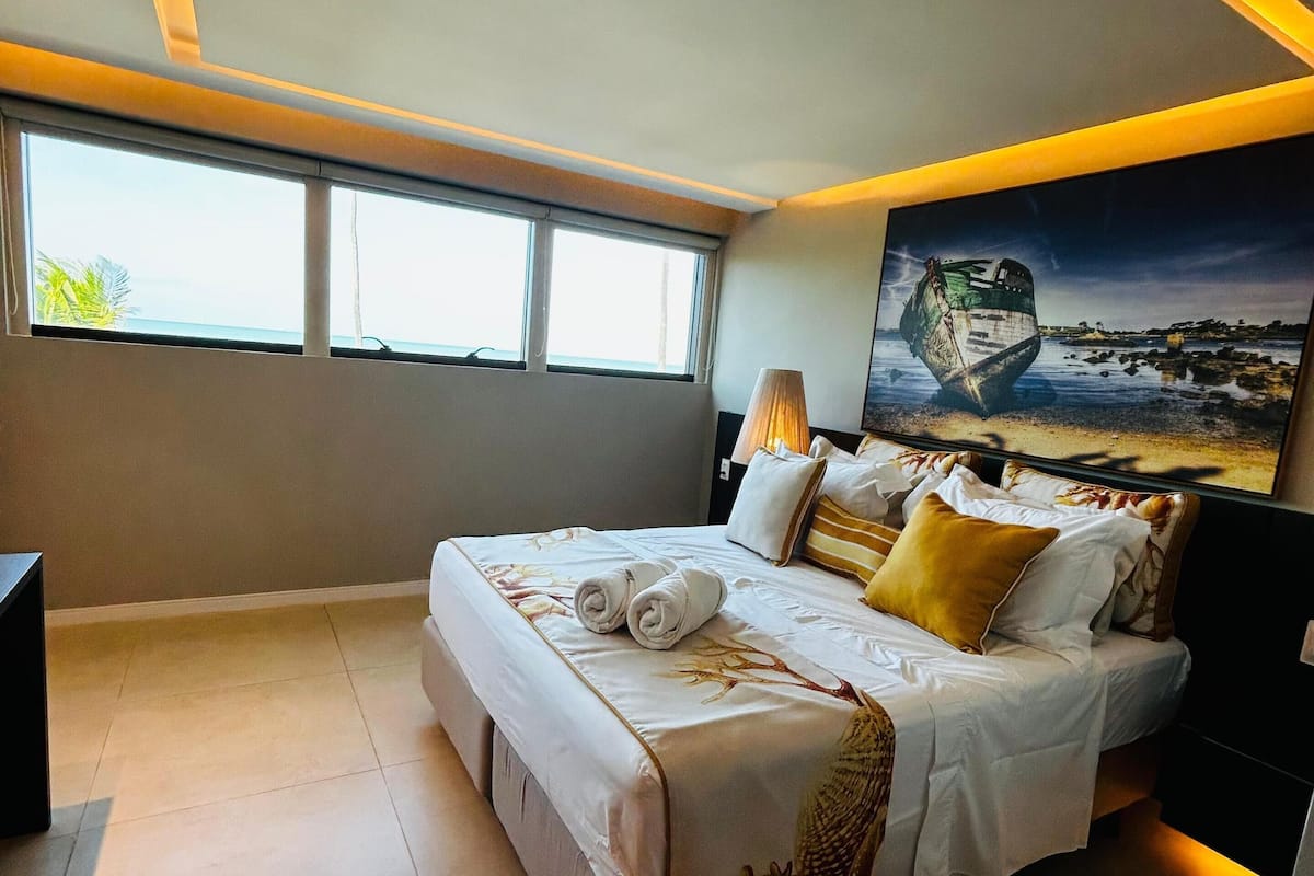 Ein modernes Schlafzimmer mit einem großen Bett, einem Gemälde eines Bootes und Meerblick durch das Fenster.