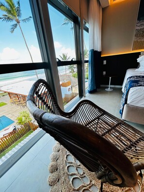 Ein Zimmer mit großem Fenster, das Blick auf Strand und Pool bietet, ein Bett mit blau-weiß gestreifter Bettdecke und ein bequemer Holzliegestuhl.