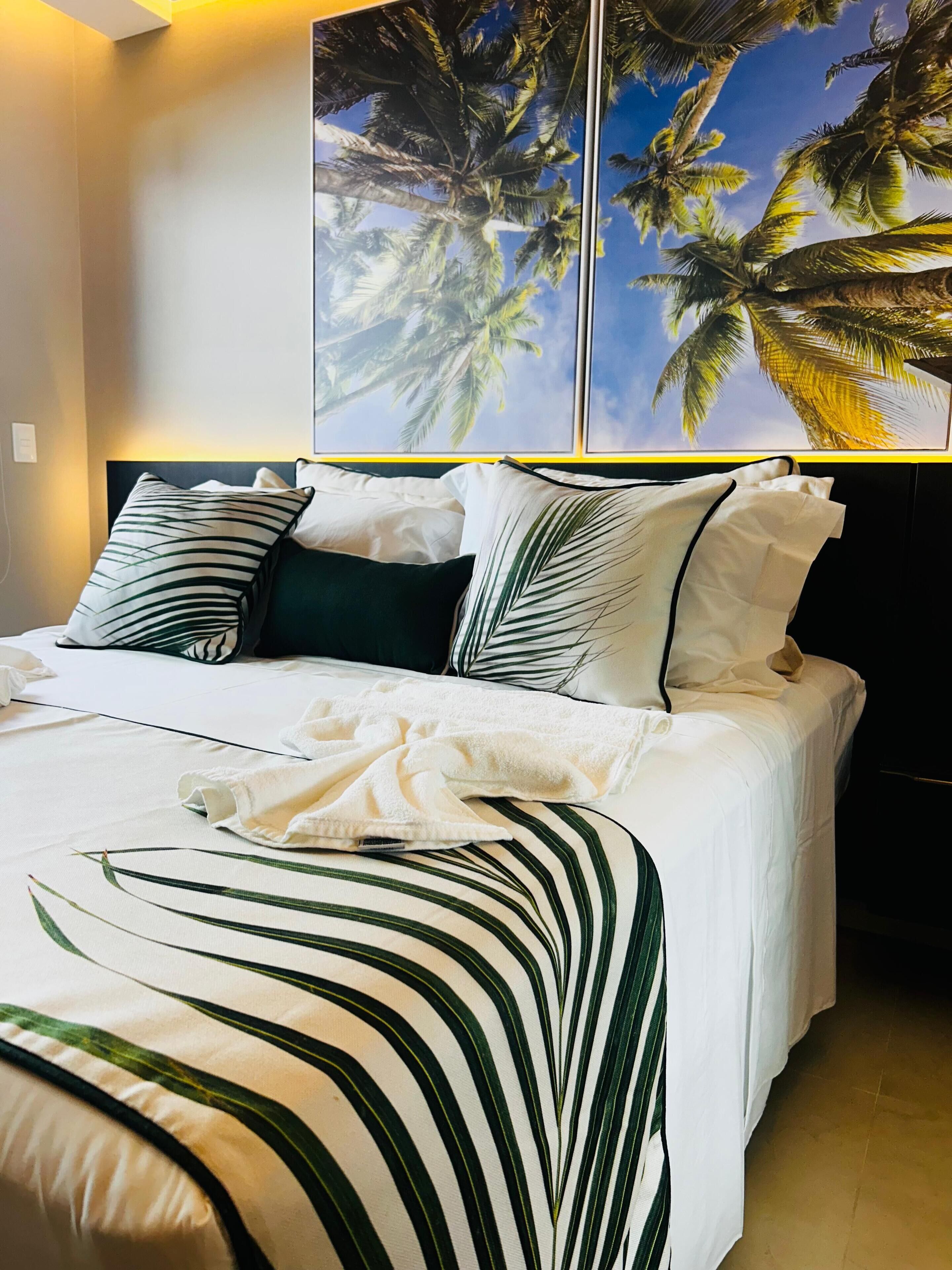 A neatly made bed with white bedding and green striped pillows, a large window with a view of palm trees, and a wall-mounted painting of palm trees.