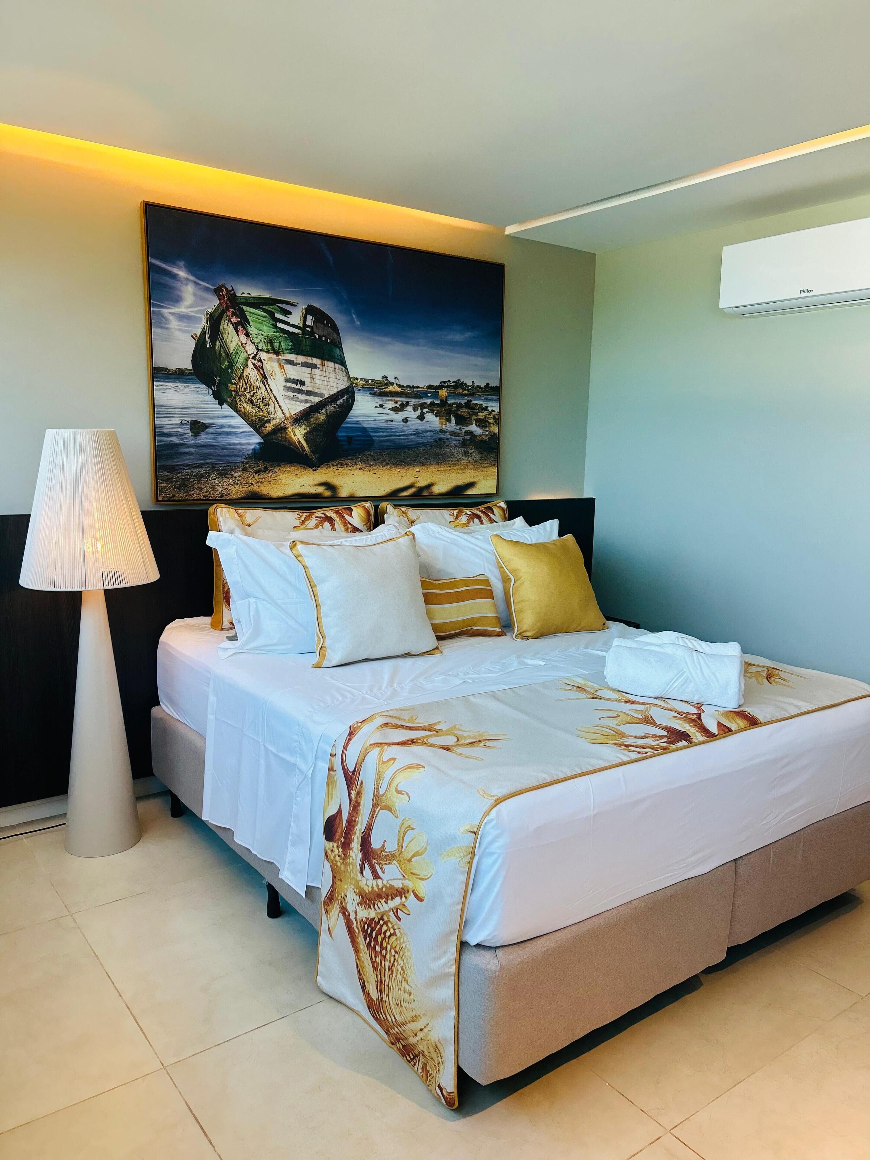 A neatly made bed with a floral bedspread, a large painting of a boat on the wall, and a floor lamp.