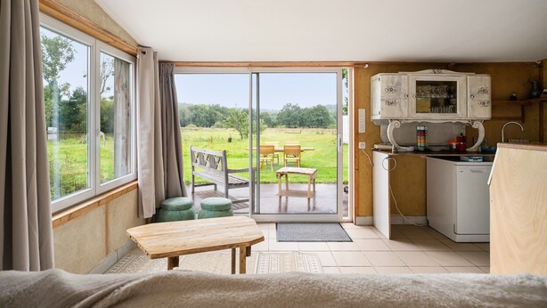 A room with a sliding glass door leading to an outdoor area, a wooden table, a bench, and a kitchenette with a cabinet. - Holiday Home 'Gîte Pour 4 Avec Sauna' with Shared Garden (Broons)