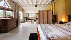 A bedroom with a large bed, a wooden dresser, a desk, and a chair. There is a window with curtains and a view of greenery outside. - Holiday Home 'Gîte Pour 4 Avec Sauna' with Shared Garden (Broons)