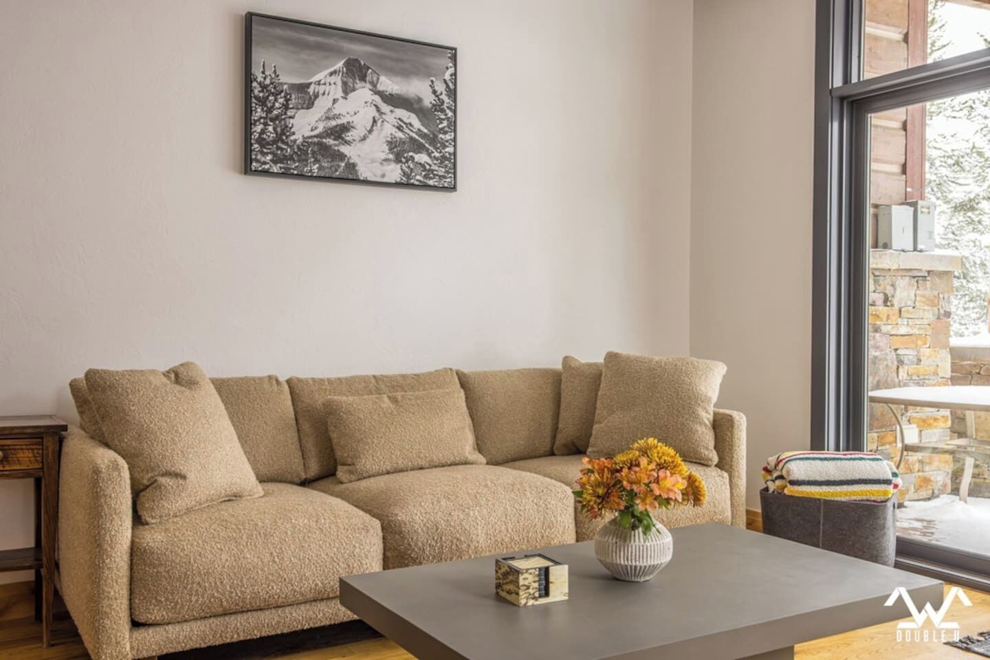 A beige sectional sofa with a vase of flowers on a table, a framed picture on the wall, and a view of a stone building through a window.