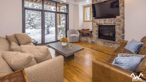Ein modernes Wohnzimmer mit Kamin aus Stein, Ledersofas, einem Flachbildfernseher und großen Fenstern mit Blick auf Schnee.