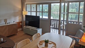 Een moderne woonkamer met een televisie, eettafel, stoelen en uitzicht op een meer door grote ramen.