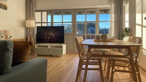 Een woonkamer met een televisie, een houten eettafel, rieten stoelen en uitzicht op bomen door grote ramen.