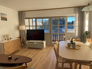 A cozy living room with a flat-screen TV, wooden furniture, a dining table with chairs, and a view of a snowy landscape through large windows. - Appartement am See (Haidmühle)