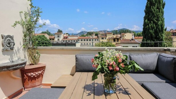 Una terraza en la azotea con una mesa de madera, cojines grises y un florero.