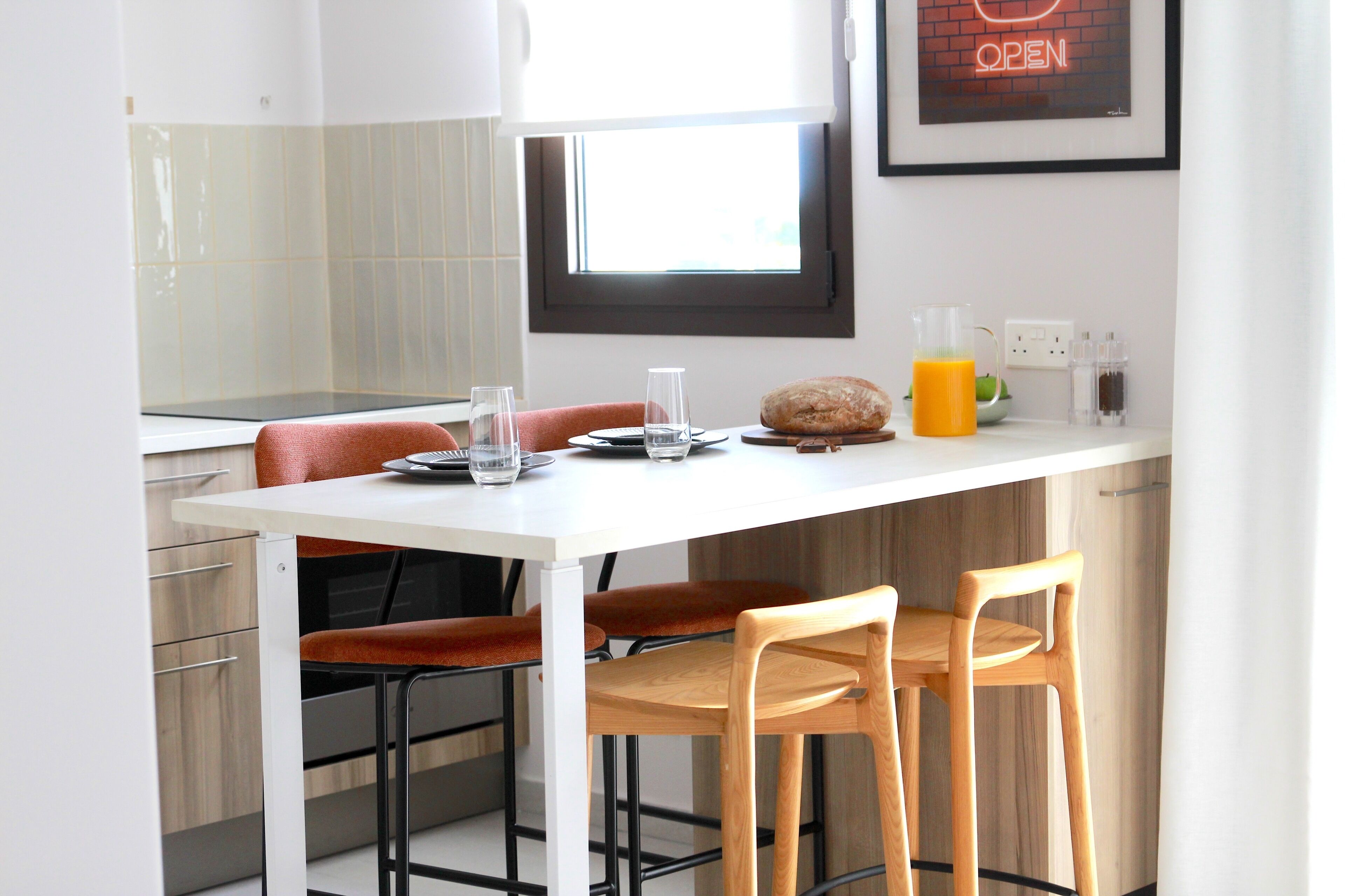 Eine moderne Küche mit einem weißen Tisch, zwei Holzstühlen, einem Brotlaib, einem Glas Orangensaft und einem Fenster mit einer Neonreklame.