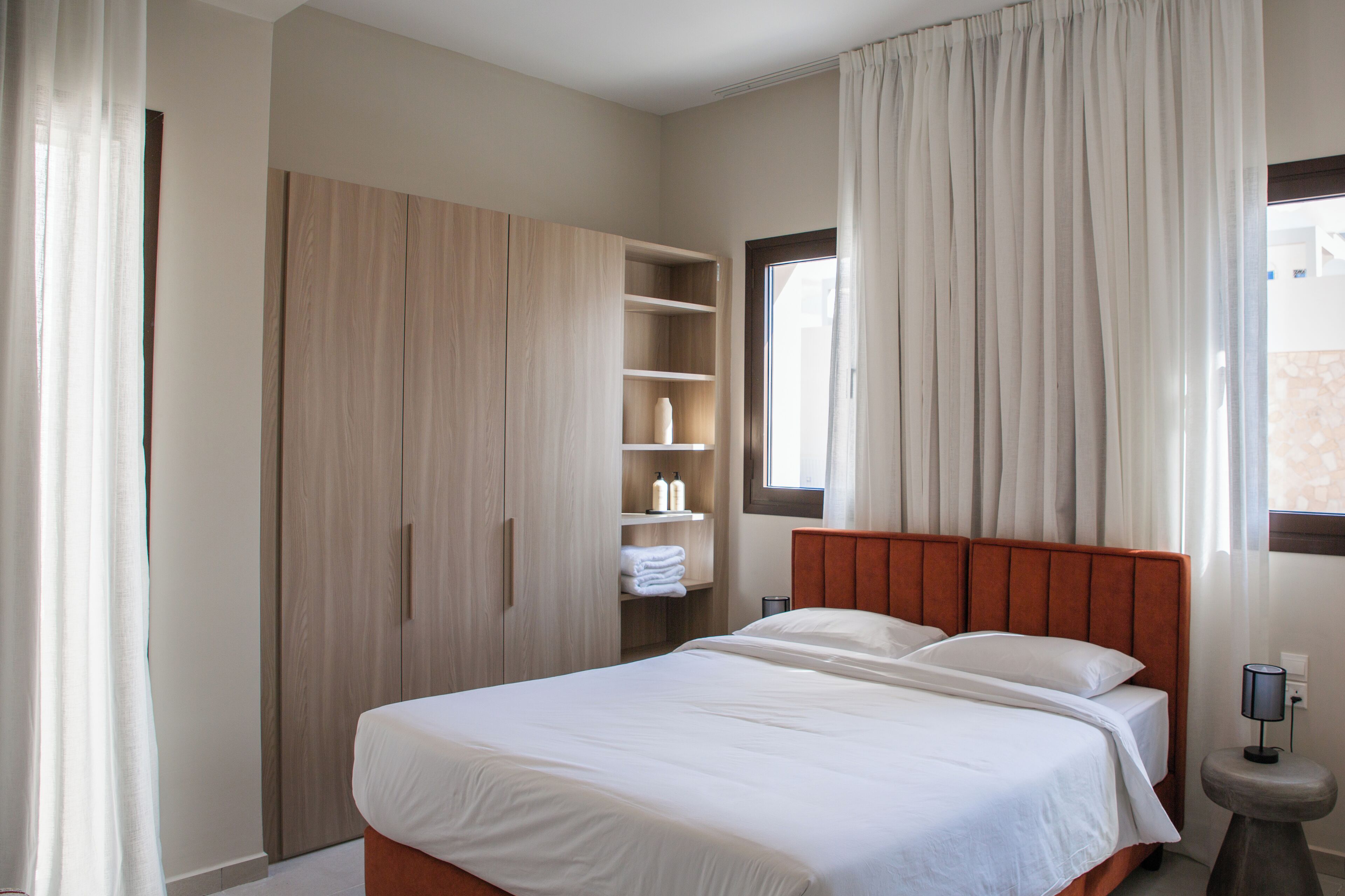 A bedroom with a bed, wooden wardrobe, shelves with toiletries, and a window with curtains.
