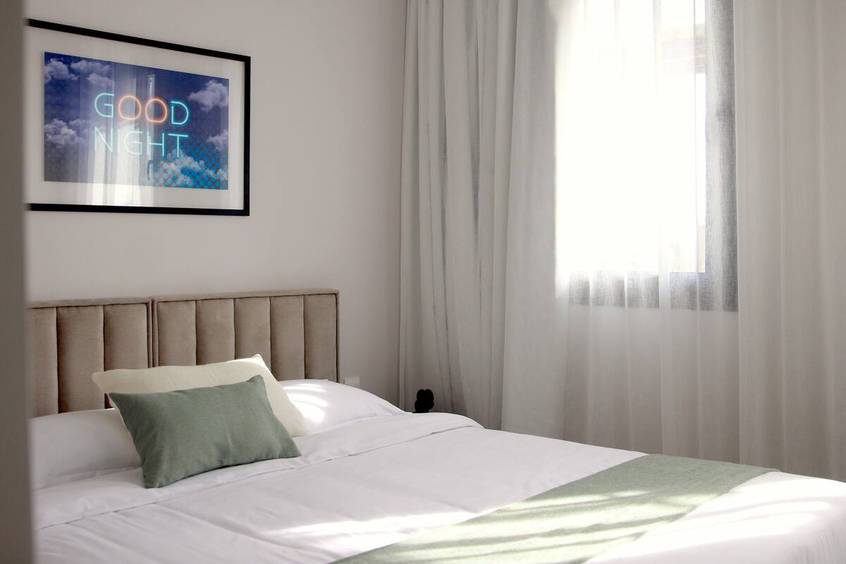 Ein ordentlich bezogenes Bett mit Kopfteil, Kissen und Decke. An der Wand hängt ein gerahmtes Bild und ein Fenster mit Vorhängen.