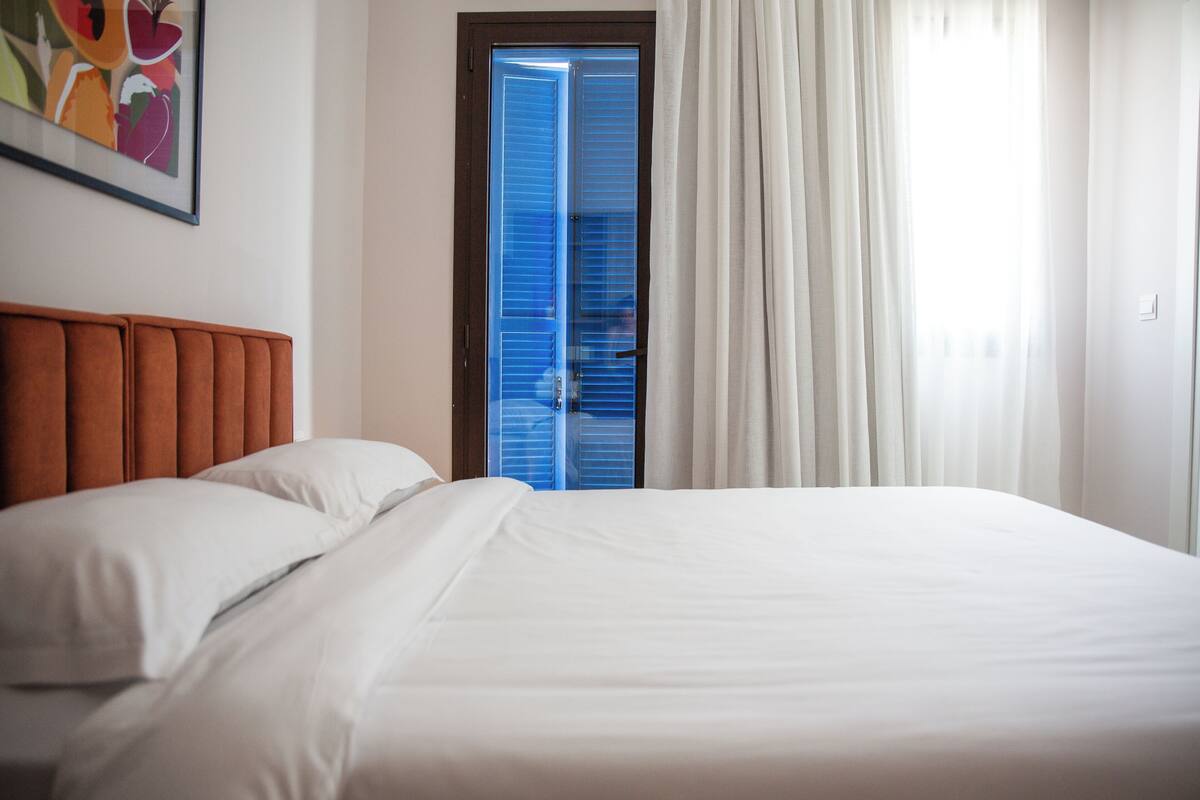 Ein Hotelzimmer mit Bett, Kopfteil, einer Tür mit blauen Fensterläden und einem gerahmten Bild an der Wand.