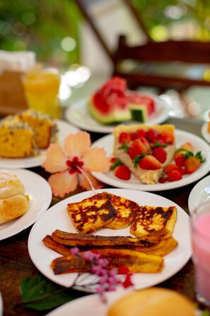 A table with various food items including a slice of watermelon, a plate of pancakes, and a glass of orange juice. - Pousada Beach Garden (Ipojuca)