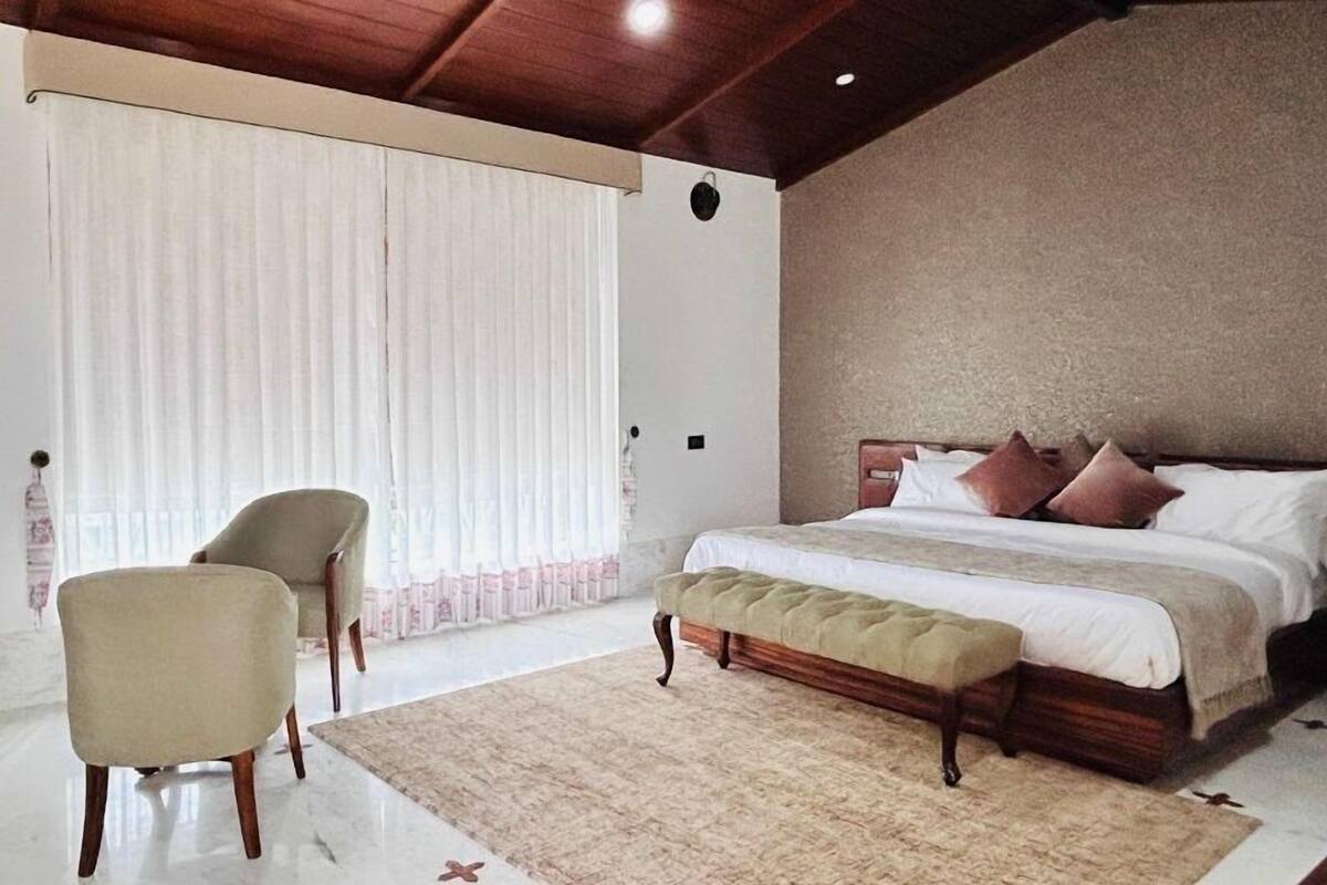 A bedroom with a bed, a chair, a bench, and a large window with curtains.