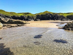 A beach with clear shallow waters, rocky terrain, and green hills in the background.
